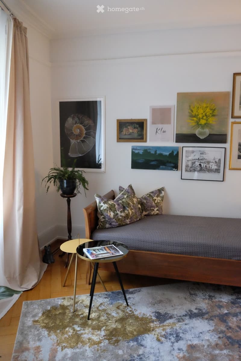 Cozy living room with a wooden sofa, patterned cushions, a small coffee table, a potted plant, and several picture frames on the wall.