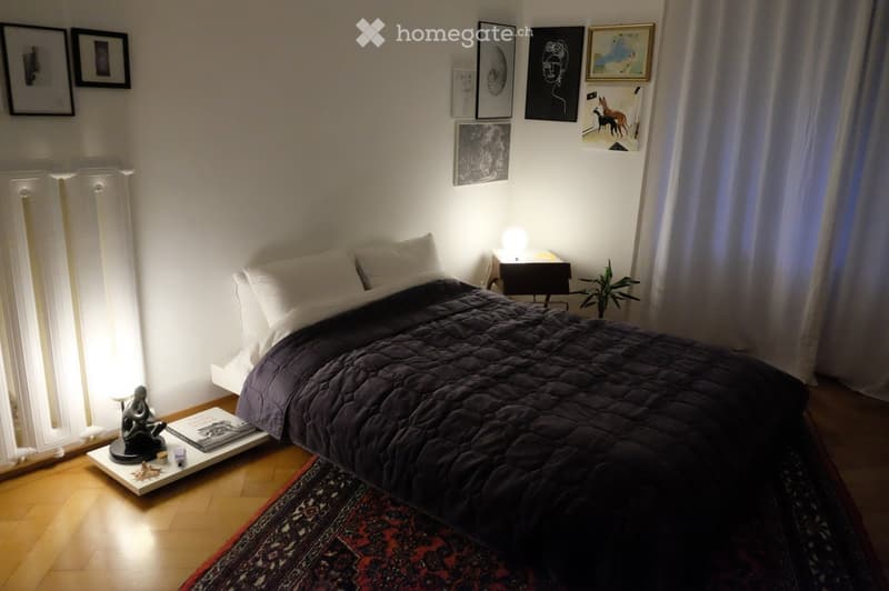 Cozy bedroom with a dark quilted bed, white pillows, a nightstand with a table lamp, a side table with books and a plant, and framed artwork on the walls.
