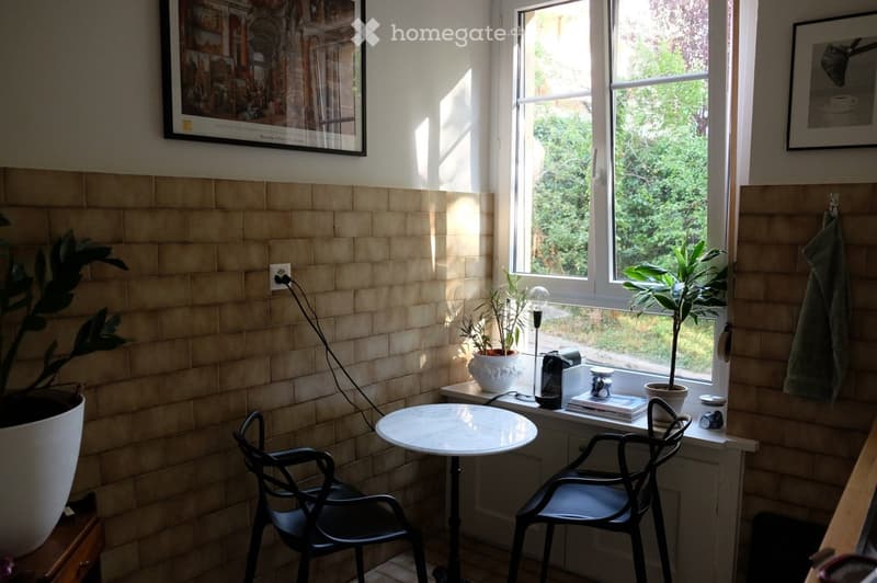 Bright room with large window, tiled walls, small round table with two chairs, potted plants, lamp, coffee maker, and framed pictures on the wall.