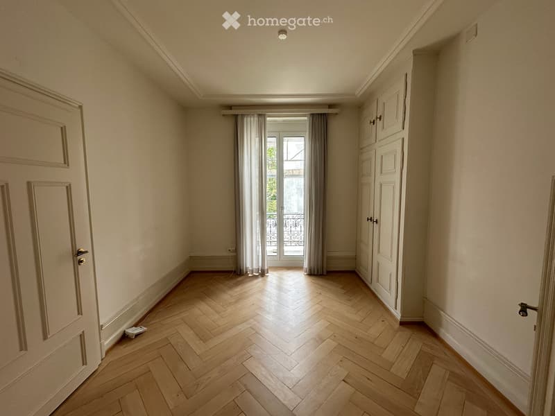 Empty room with wooden herringbone flooring, white walls, and a large window with curtains leading to a balcony.