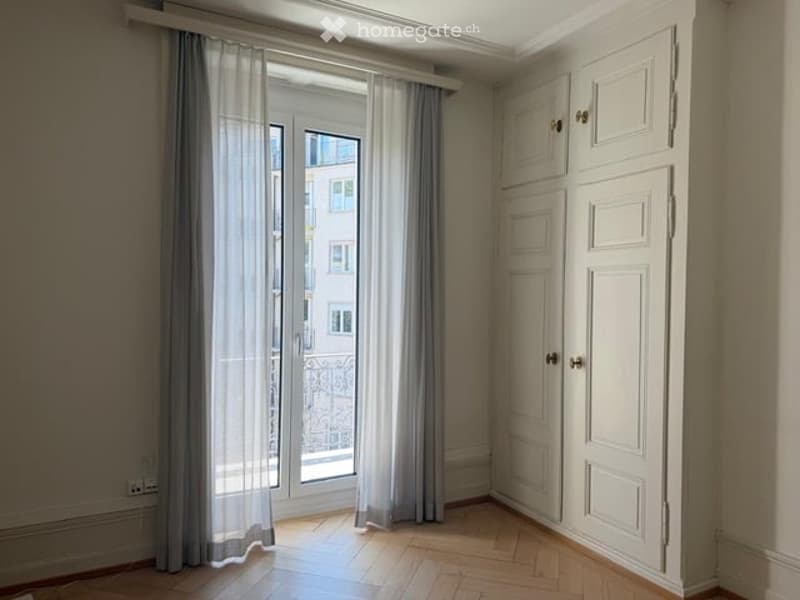 Empty room with wooden flooring, large window with grey curtains, white built-in cabinetry, and a view of a building outside.