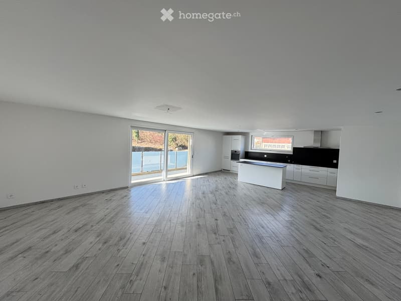 Open-plan living and dining area with wooden flooring, white walls, and large sliding glass doors leading to a balcony. Kitchen area with white cabinets and a black countertop is visible in the background.