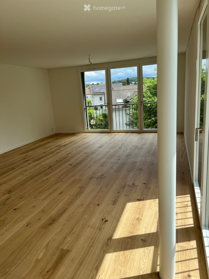 Empty room with light wooden flooring and large sliding glass doors leading to a balcony with a view of the surrounding area.