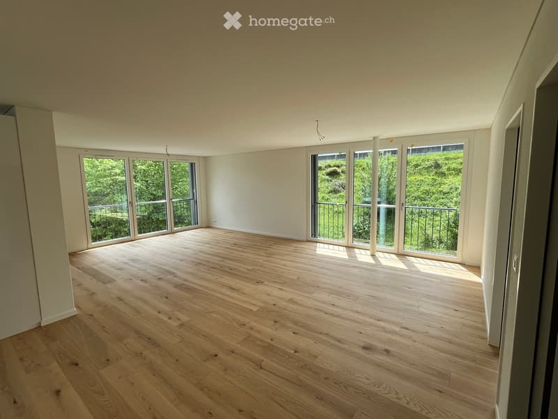 Empty room with light wooden flooring and large glass doors leading to a balcony with a view of greenery.