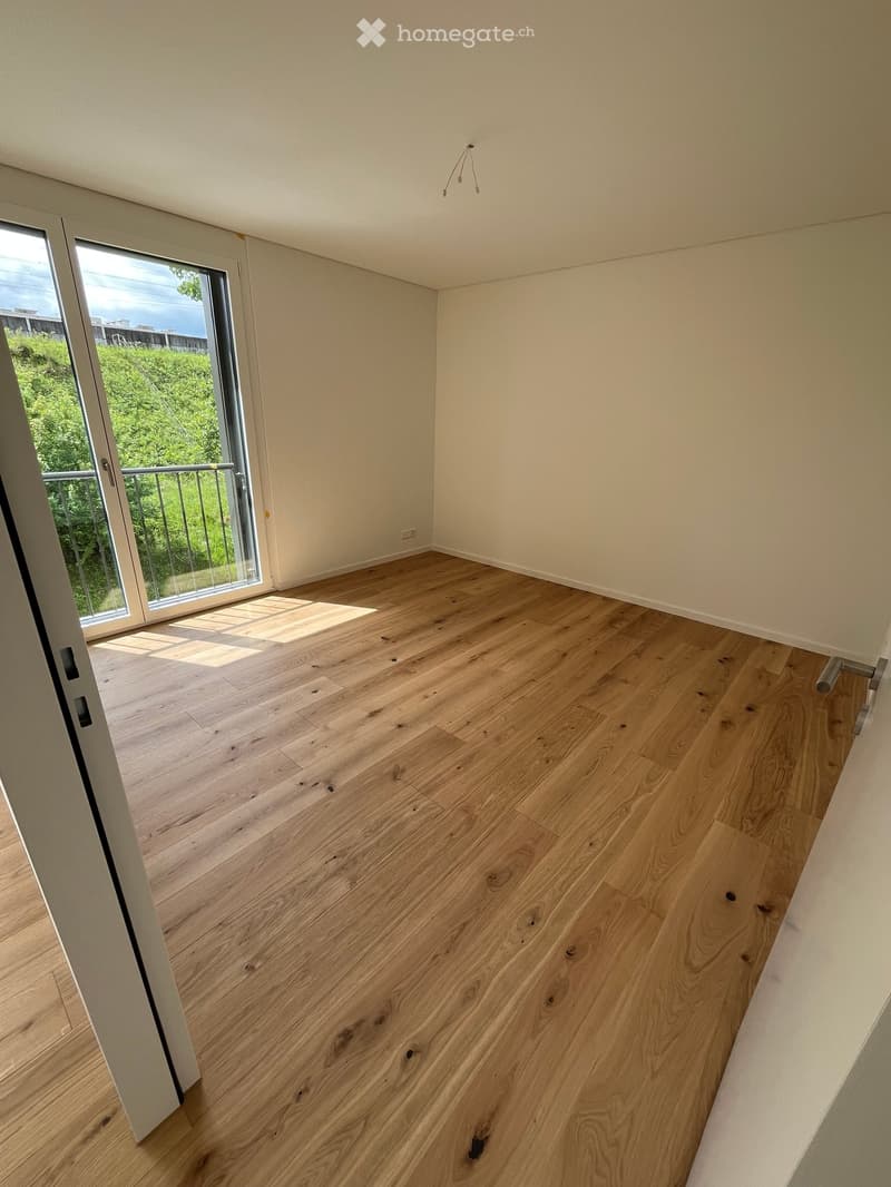 Empty room with light wooden flooring and white walls, featuring a large glass door leading to a balcony with a view of greenery.