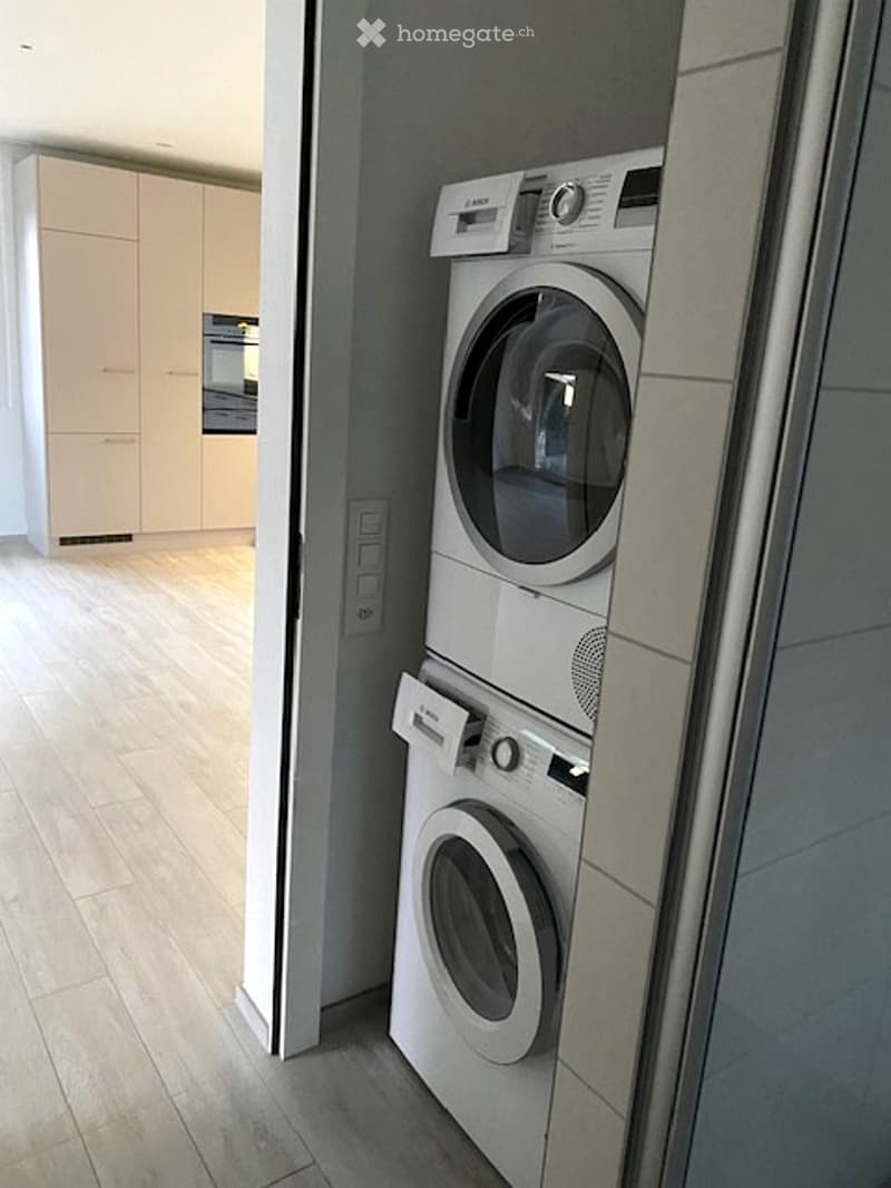 Modern bathroom with a stacked washing machine and dryer unit, light wood flooring, and light-colored cabinetry visible in the background.