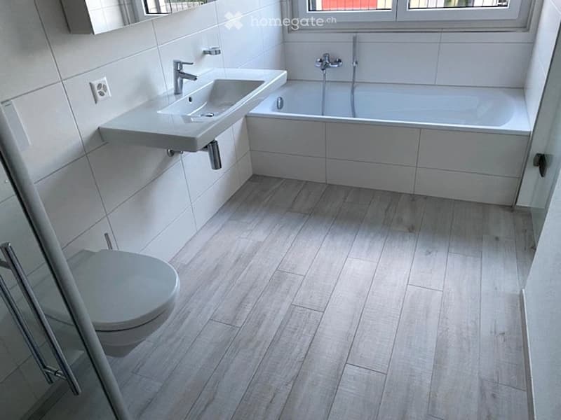 Modern bathroom with white tiles, a floating sink, a bathtub, and a toilet. The floor is covered with light wood tiles, and the walls are white.