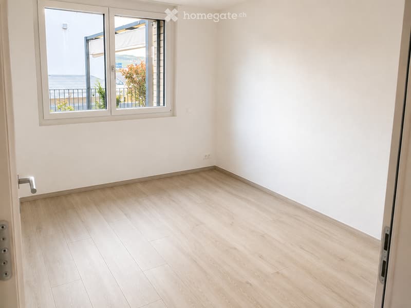 Empty room with light wooden flooring, white walls, and a large window with a view of a balcony and outdoor area.