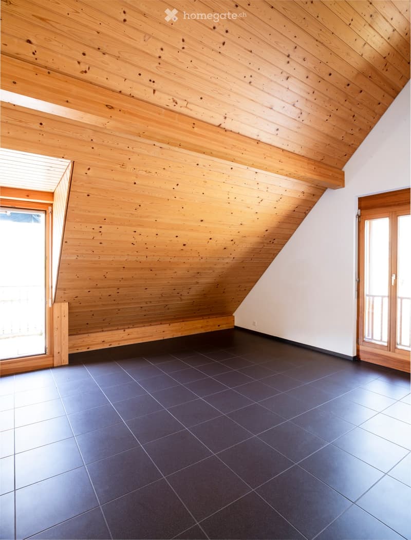An empty attic room with wooden walls and ceiling, dark tiled floor, and large windows allowing natural light.