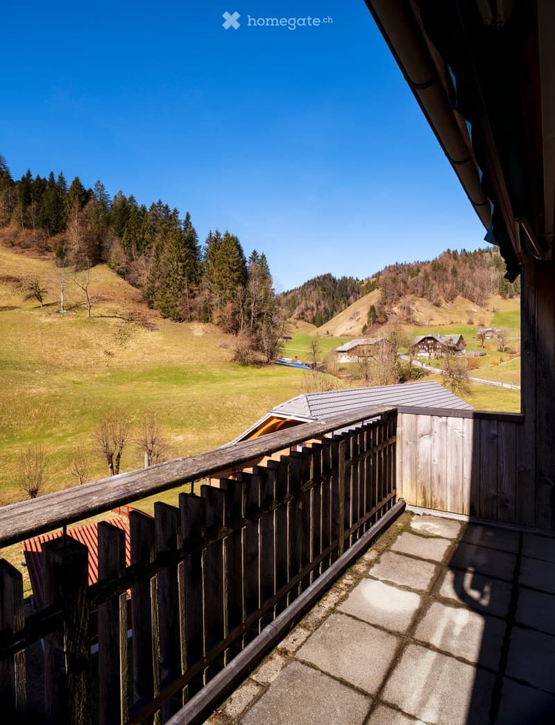 Balcony with wooden railing overlooking a scenic countryside with green fields, scattered trees, and distant houses.