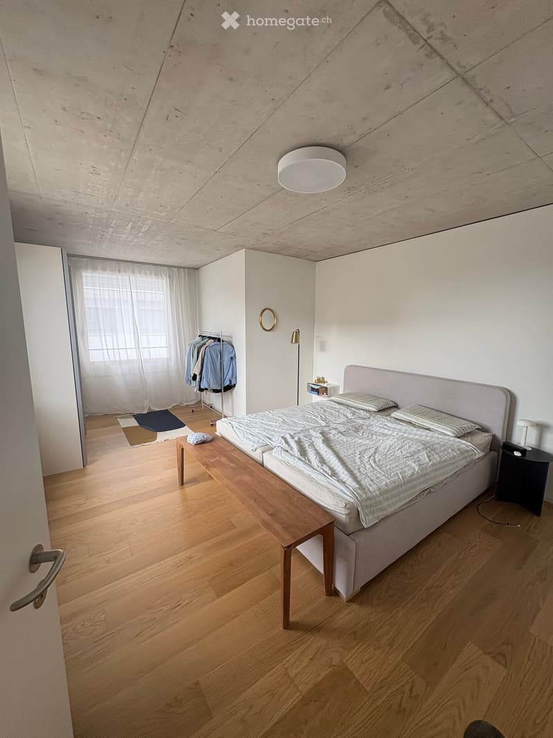 Modern bedroom with wooden flooring, a large bed with a headboard, a nightstand, a small table, a clothing rack, and a large window with sheer curtains.