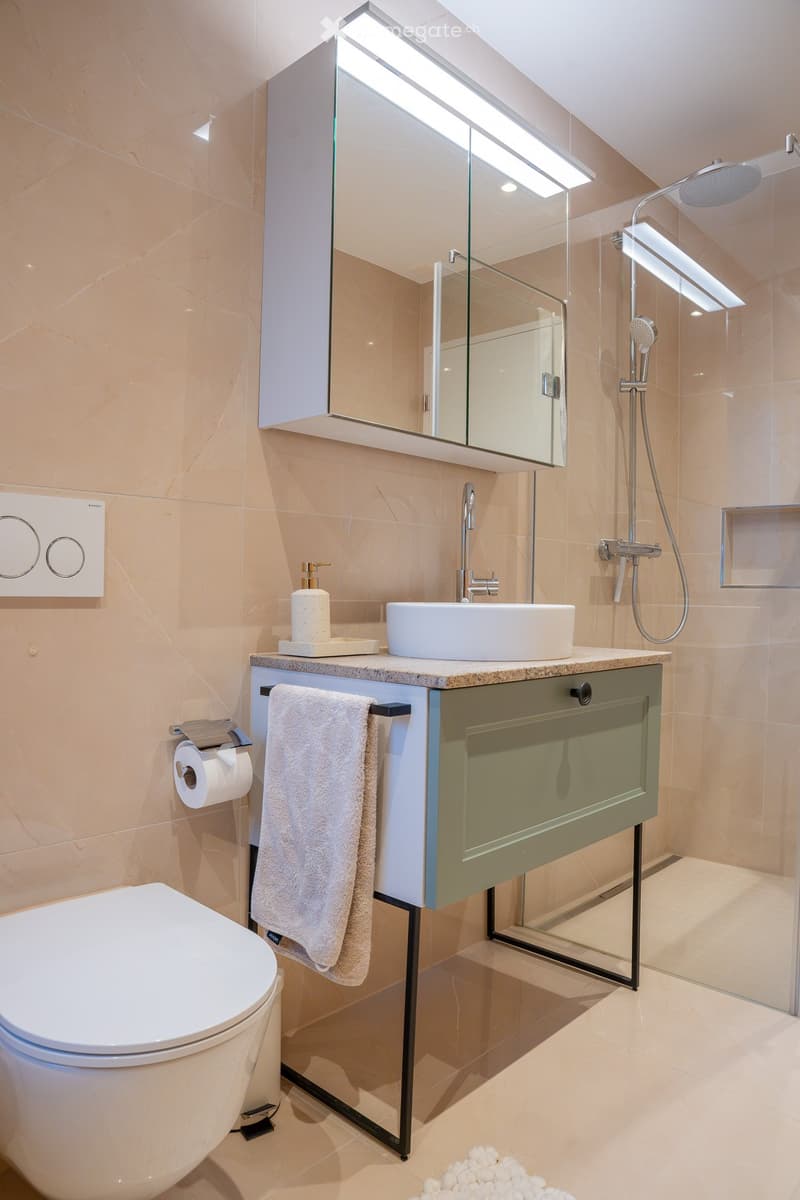 Modern bathroom with beige tiles, a white toilet, a sink with a modern faucet, a large mirror, a towel, and a glass-enclosed shower area.
