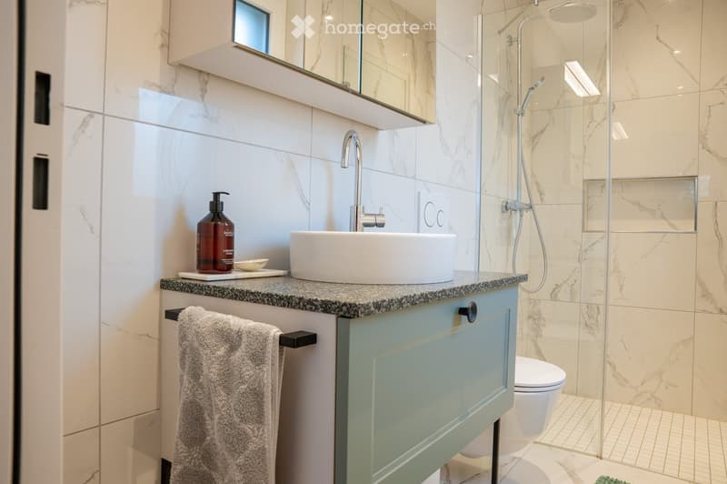 Modern bathroom with marble tiles, a white sink, a glass-enclosed shower, and a toilet. A towel hangs on a rack, and a bottle is placed on the countertop.