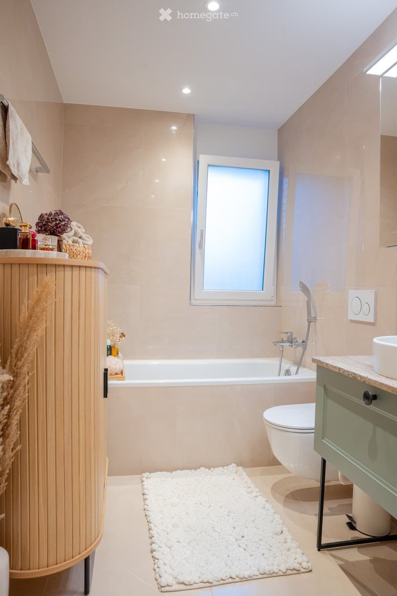 Modern bathroom with beige tiles, a bathtub, toilet, sink, and a wooden cabinet. Features a white pebble rug, a towel, and decorative flowers on the cabinet.