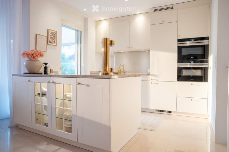 Modern kitchen with white cabinets, a built-in oven, a central island with glass-fronted cabinets, and a vase with flowers on the counter.