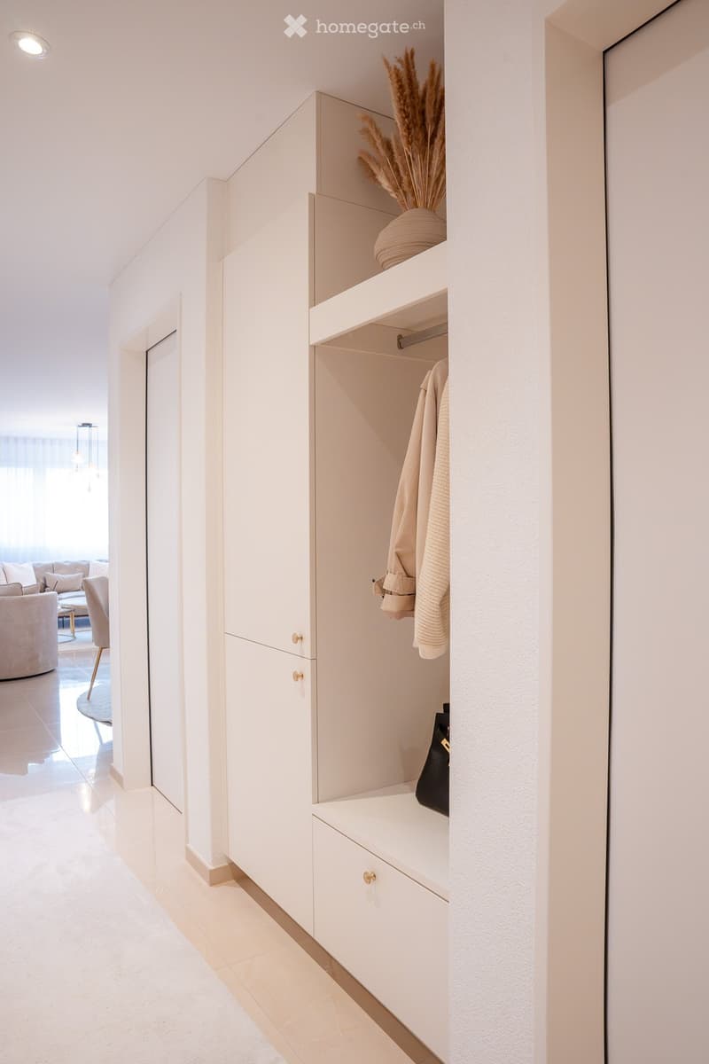 Modern hallway with white built-in storage unit, open shelving, and a small bench. Visible items include a vase with dried flowers, folded towels, and a handbag. The hallway leads to a living area with a sofa and coffee table.