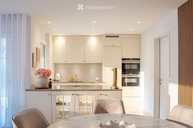 Modern kitchen with white cabinets, marble countertops, built-in ovens, and a dining area with a marble table and chairs.