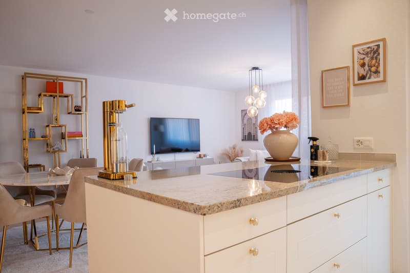 Modern kitchen with a marble countertop, white cabinets, a dining table with chairs, a television, a decorative vase with flowers, and a sleek gold shelf unit.