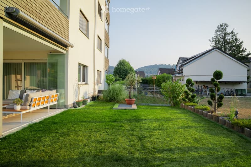 Well-maintained garden with a neatly trimmed lawn, potted plants, and a modern bench on a patio area adjacent to a house with large windows and a balcony.