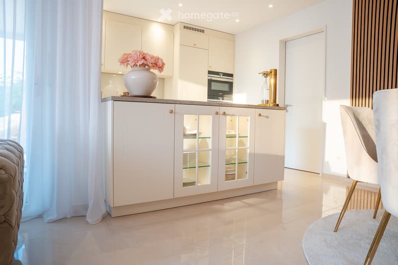 Modern, minimalist room with white cabinets, a sleek countertop, a vase with pink flowers, and a white chair with gold legs.