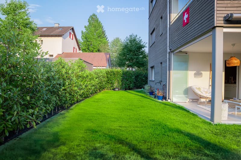 Well-maintained garden with a neatly trimmed lawn, surrounded by a hedge and trees. A modern house with a balcony and outdoor seating area is visible in the background.