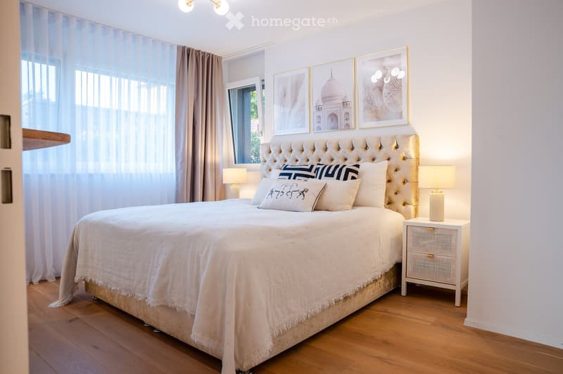 Elegant bedroom with a large tufted headboard, white bedding, decorative pillows, nightstand with lamp, and large windows with curtains.
