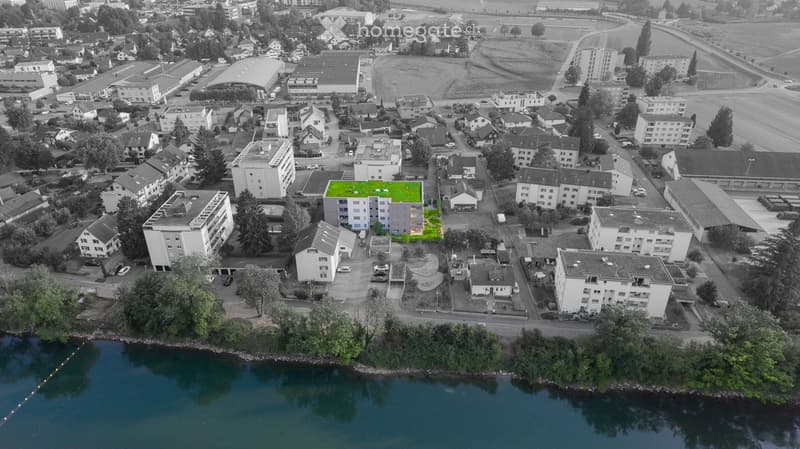 Aerial view of a residential area with multiple apartment buildings, parking lots, and a small green lawn area in the center. The area is bordered by a river on one side and surrounded by trees and open fields.