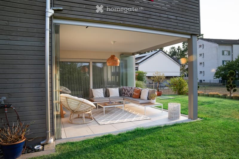 Modern outdoor patio with a sofa, coffee table, wicker chair, and potted plants, connected to a house with large glass doors.