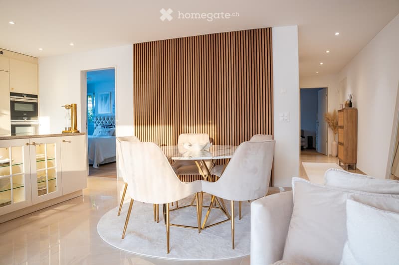 Modern open-plan living and dining area with white dining chairs, a glass table, a sleek kitchen with cabinets and an oven, and a visible bedroom with a tufted headboard and pillows.