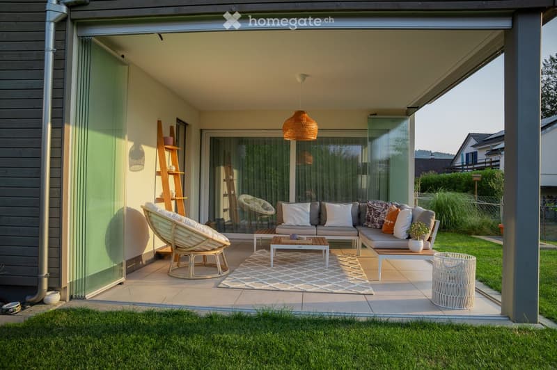 Modern covered outdoor space with a sofa, coffee table, wicker chairs, and a potted plant, connected to the house via large glass doors.