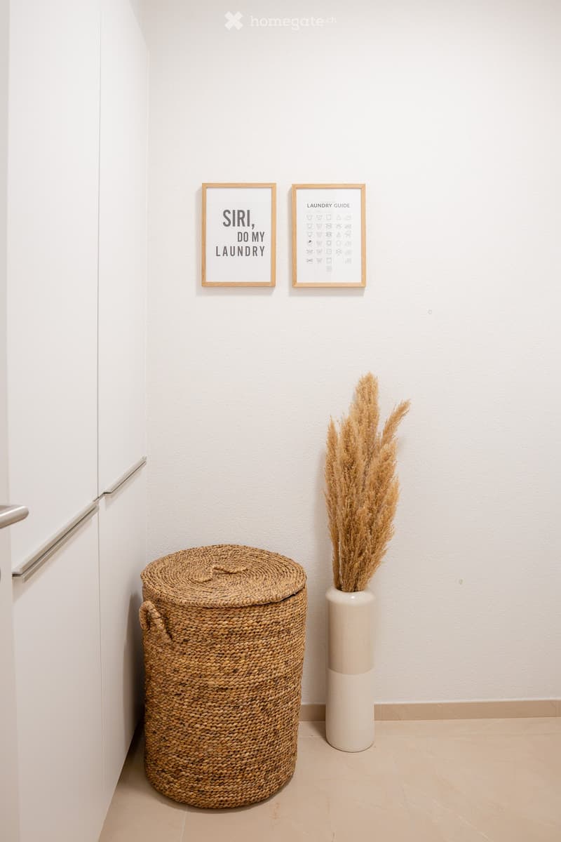 A hallway or entryway with a woven wicker basket, a tall vase with dried pampas grass, and two framed prints on the wall.