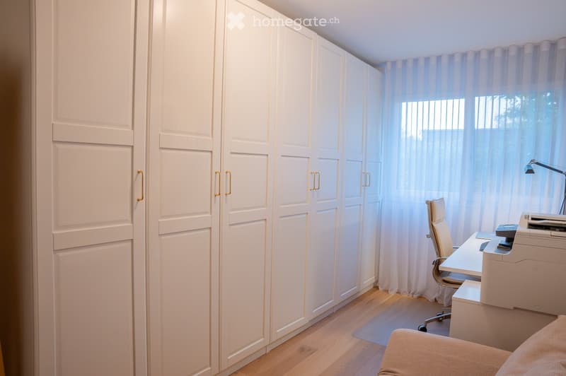 A well-lit room with a large white cabinet, a desk with a chair, a printer, and a window with sheer curtains.