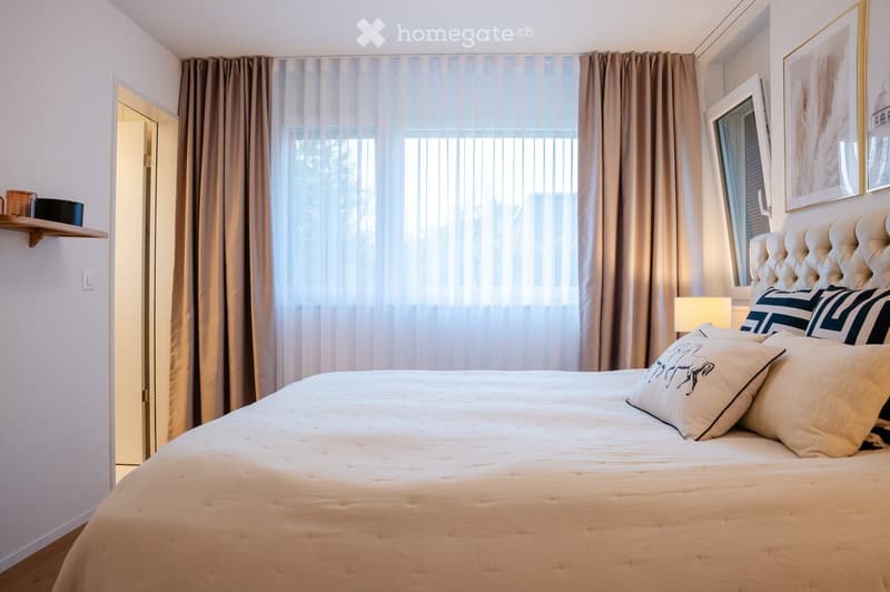 Elegant bedroom with a large, tufted bed, decorative pillows, a nightstand with a lamp, and a window with sheer curtains. A small shelf with decorative items is visible on the left side.