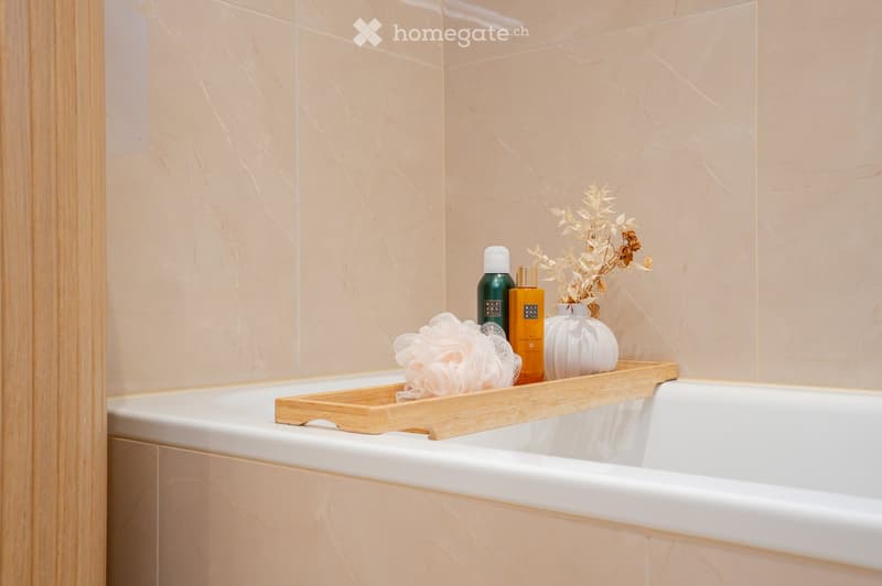 A modern bathroom with a white bathtub, a wooden tray holding bottles, a vase with dried flowers, and a sponge. The walls are tiled in a light beige marble pattern.