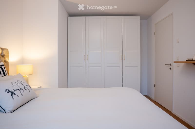Modern bedroom with a large bed, decorative pillows, a bedside lamp, and a white wardrobe with gold handles.