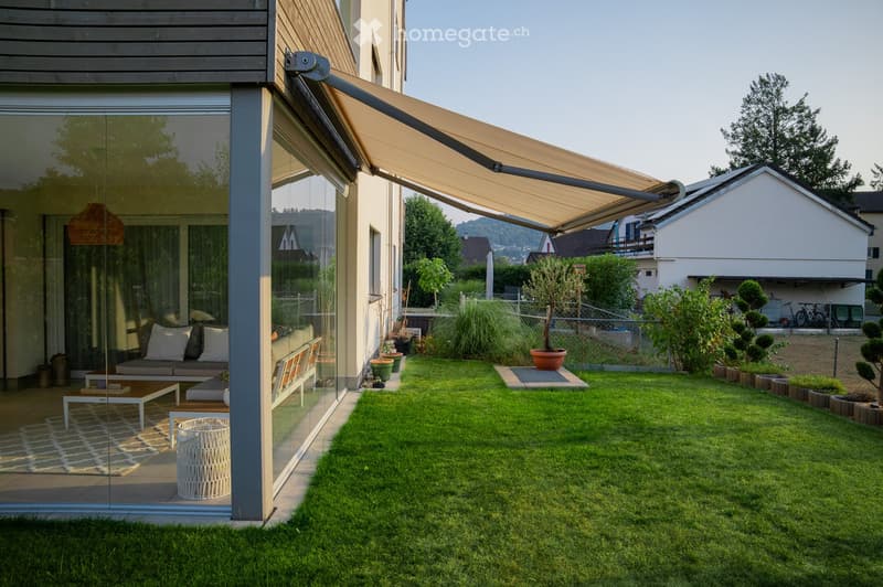 Modern outdoor patio area with a glass-walled extension, featuring a sofa, coffee table, and wicker basket. The garden includes a well-maintained lawn, potted plants, and a retractable awning providing shade.