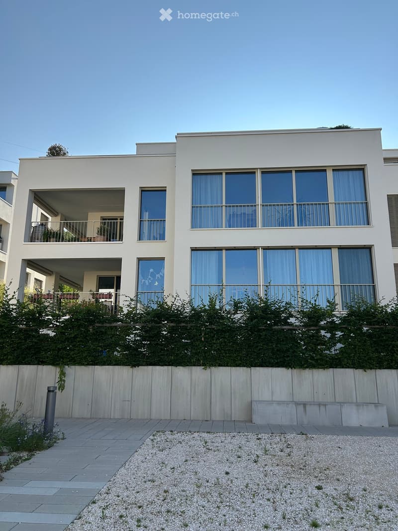 Modern two-story house with large windows, balconies, and a low hedge in front.