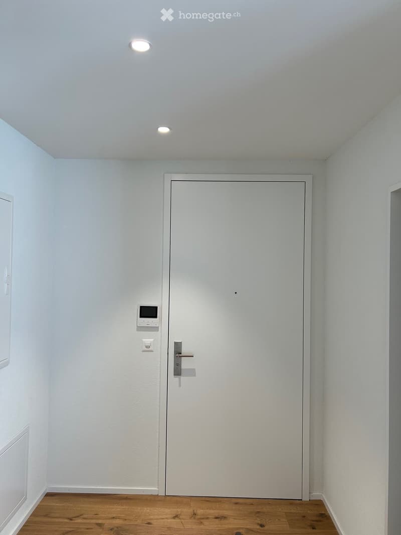 Empty room with white walls, wooden floor, and a closed white door with a silver handle. There is a small digital display and a light switch on the wall next to the door.