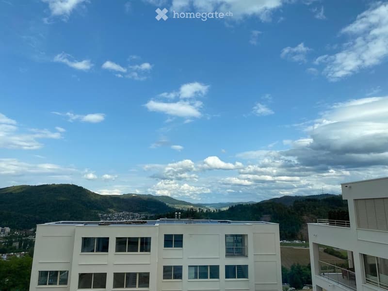 Modern white building with multiple windows and balconies, overlooking a scenic landscape with hills and a partly cloudy sky.