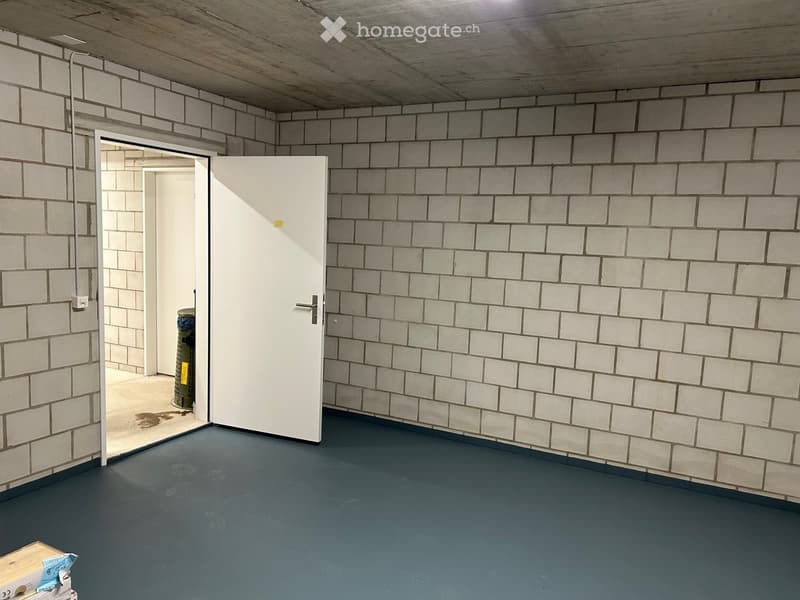 Empty room with grey tiled walls, grey concrete floor, and a white door leading to another space.