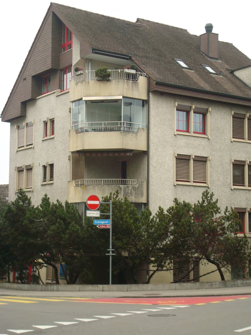 Two-story residential building with balconies, red-framed windows, and a tiled roof. Features include a stop sign and street signs in front.
