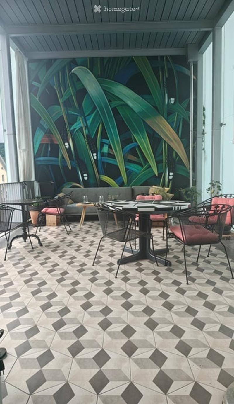 A modern, stylish outdoor seating area with a geometric tile floor, a large leafy mural backdrop, and contemporary furniture including a sofa, tables, and chairs.