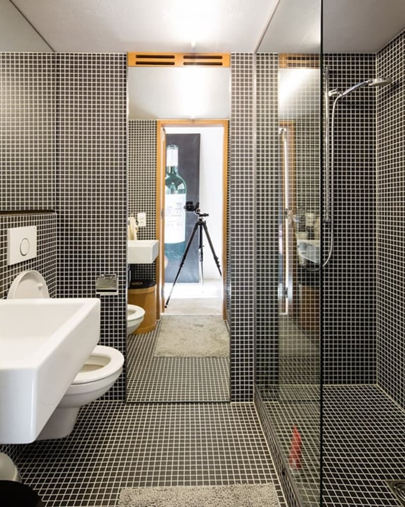 Modern bathroom with black and white tiled walls and floor, white toilet, sink, and glass-enclosed shower. A camera on a tripod is positioned in the center of the room.