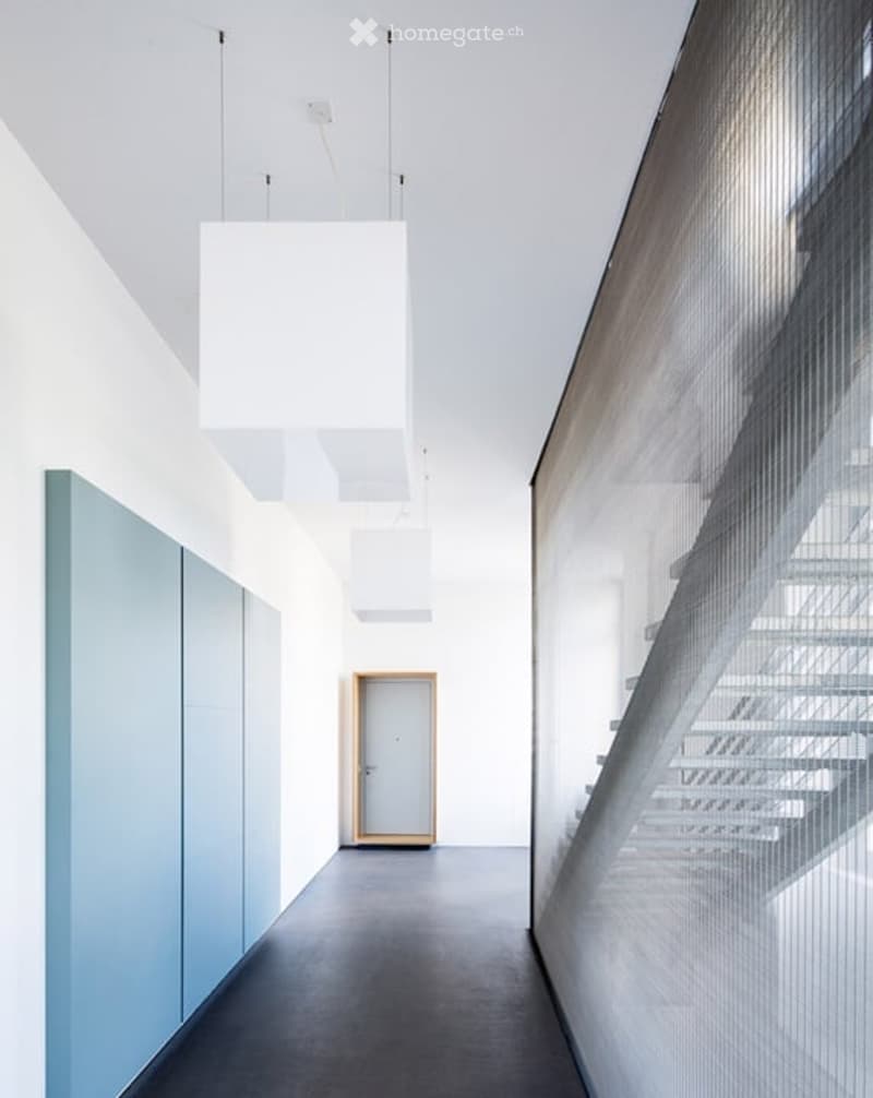 Modern hallway with white walls, grey carpeted floor, a single door, and a staircase visible through a glass wall. Minimalist design with a pendant light fixture hanging from the ceiling.