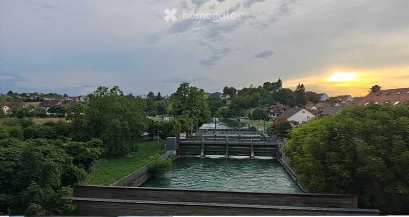 View of a dam and water channel surrounded by greenery and houses in the background, with a sunset sky.