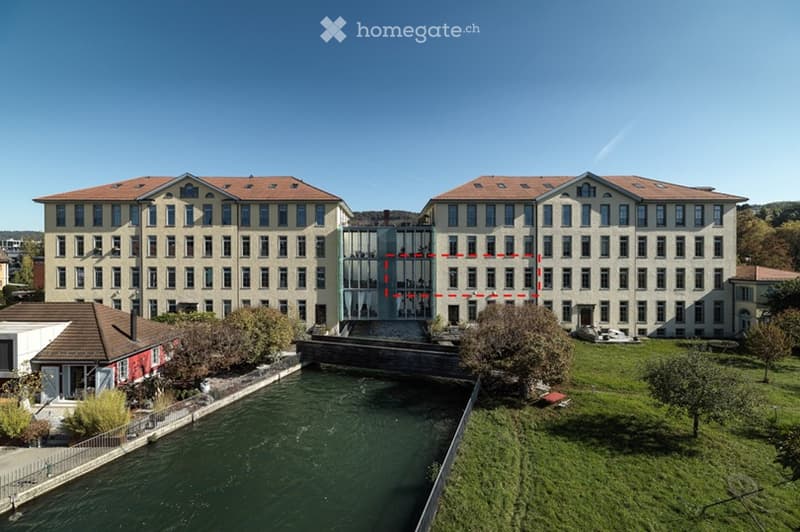 A large, multi-story industrial-style building with a symmetrical facade, numerous windows, and a modern glass extension in the center. The building is surrounded by a canal and greenery.