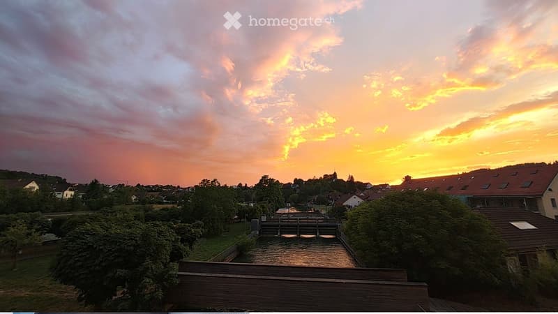 A scenic view of a small dam and surrounding landscape during sunset, with a mix of residential buildings and trees in the background.