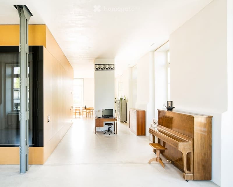 A long, bright corridor with white walls and a polished floor, featuring a wooden piano on the right, a desk with a chair in the middle, and a small dining area with tables and chairs at the far end.