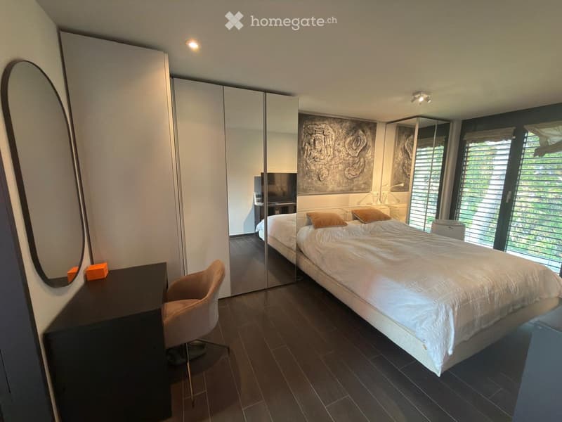 Modern bedroom with a large bed, white bedding, two throw pillows, a sleek wardrobe with mirrored doors, a desk with a chair, and a large mirror on the wall.