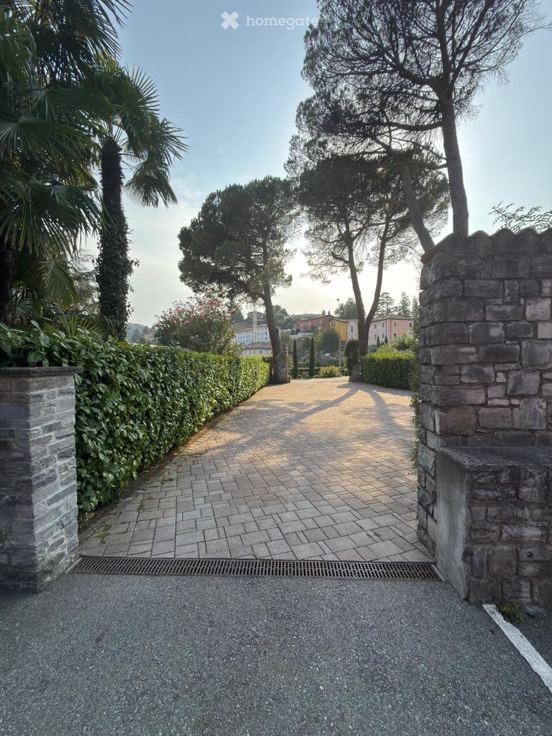 A paved pathway lined with greenery and trees leads to a gated entrance, with a stone wall on one side and a hedge on the other.